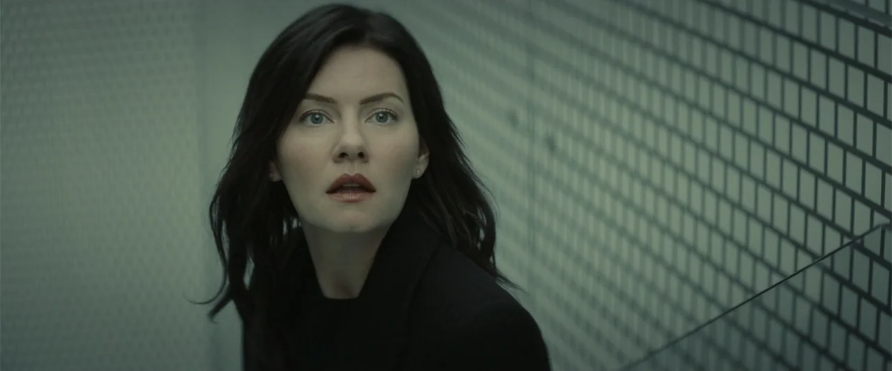 The Cellar (2022) movie still: closeup — A sterile industrial stairwell features a metallic grid wall and cool, desaturated lighting. A woman in a…