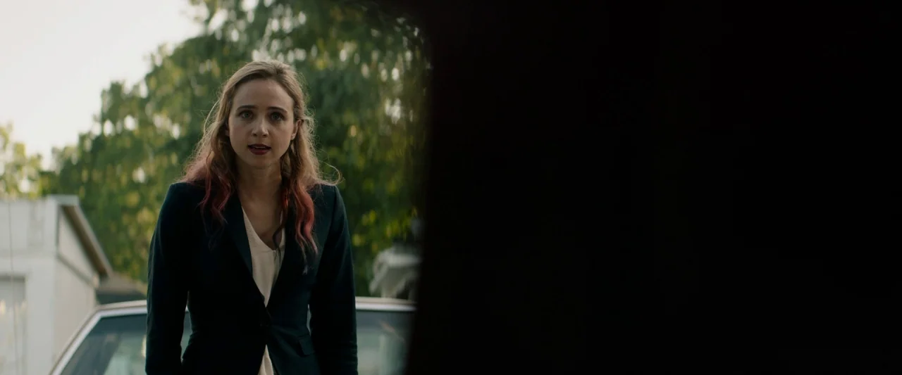 The Monster (2016) movie still: medium — A woman stands in a residential driveway with trees blurred in the background. She is captured in a dirty…