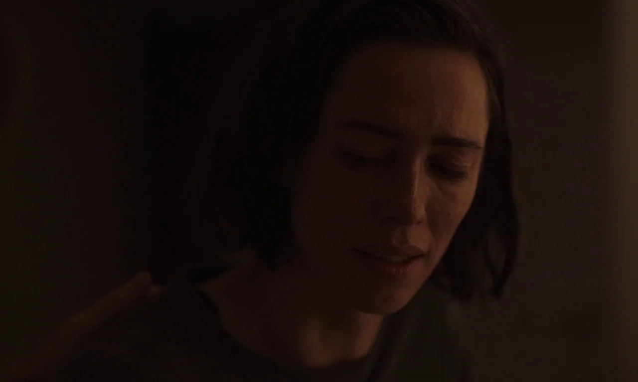 Resurrection (2022) movie still: closeup — A dimly lit, shadowed space focuses tightly on a woman's face and shoulders. A hand rests on the woman's…