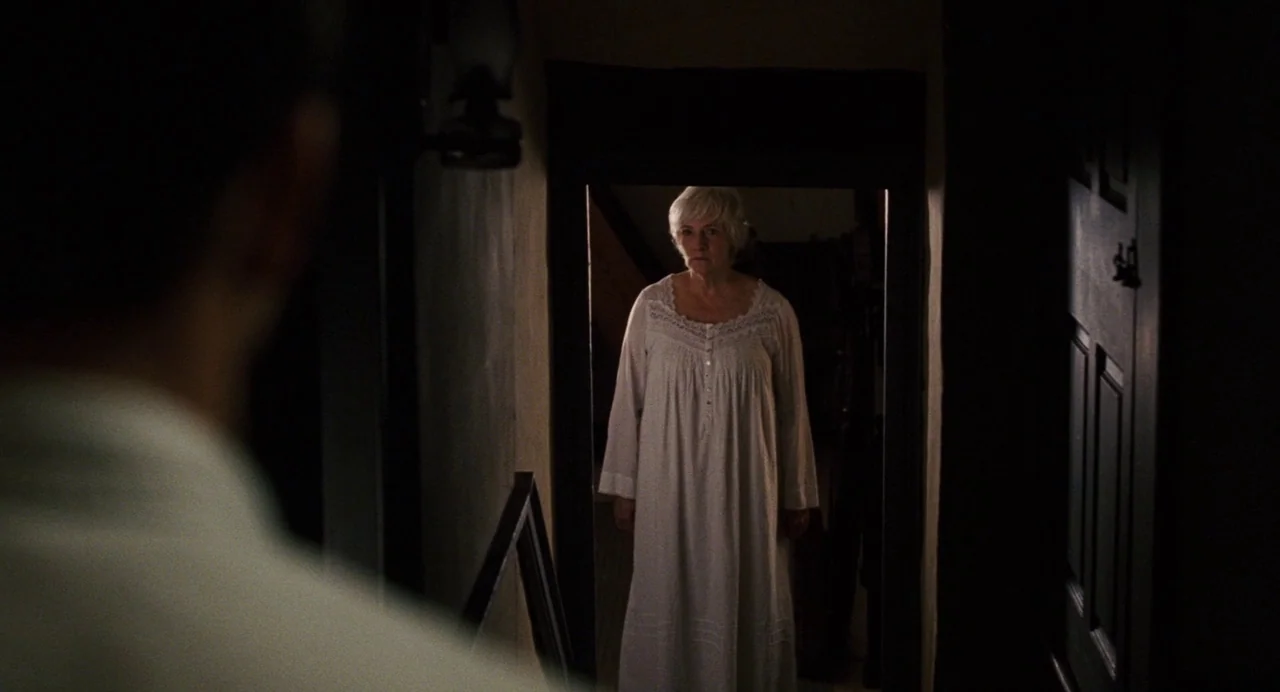 The Happening (2008) movie still: medium — An old residential hallway features dark walls and a framed doorway leading into darkness. An elderly…