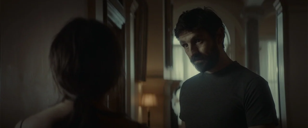 The Cellar (2022) movie still: medium — A dark, dimly lit domestic corridor features a man looking towards a woman's back. The man wearing a dark…