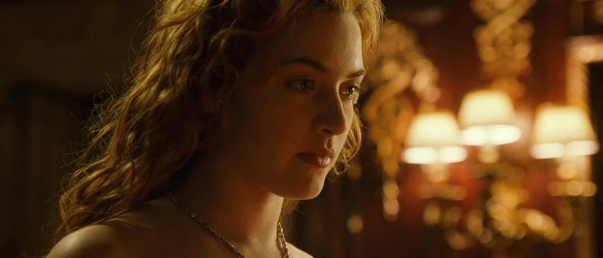 Titanic (1997) movie still: closeup — A dimly lit room with ornate, dark-toned walls and golden decorative accents. A young woman with long auburn…