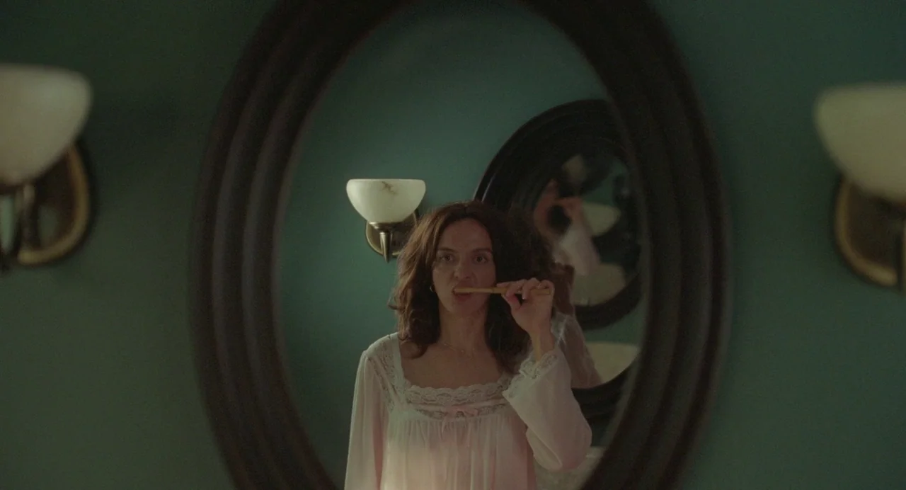 Babysitter (2022) movie still: medium — A teal-walled room contains an oval mirror mounted above a sink area. A woman in a pink lace-trimmed gown…