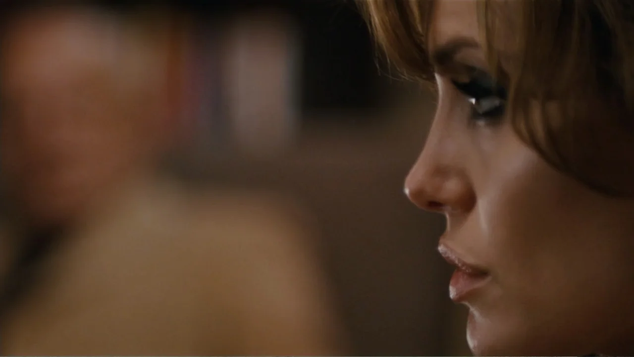 The Tourist (2010) movie still: closeup — An interior space with a blurred, out-of-focus figure in the background. A woman is seen in a close-up…
