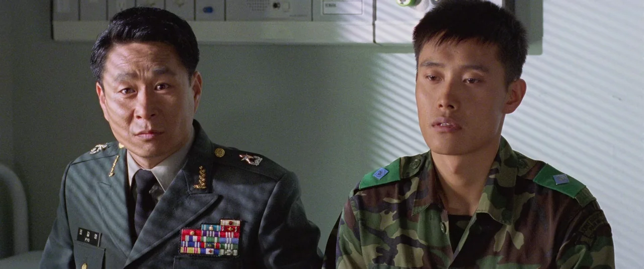Joint Security Area (2000) movie still: medium — An interior medical room features a wall-mounted medical monitor behind two soldiers. A mature…