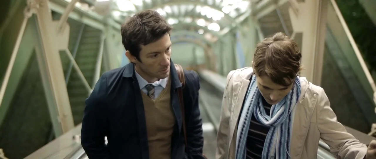 Upstream Color (2013) movie still: medium — An enclosed glass escalator structure with metal frames provides a tunnel-like passage. A man and a…