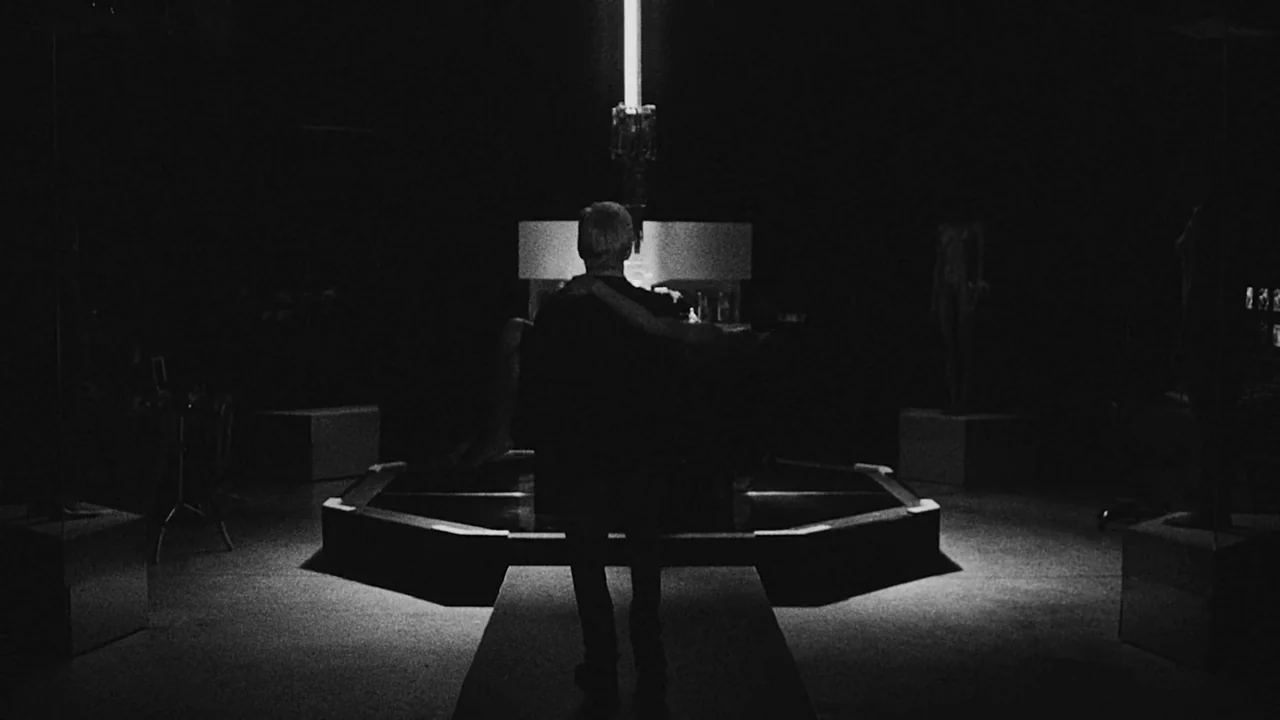 Divinity (2023) movie still: fullbody — A dark gallery space with pedestals holding statues and a single vertical light fixture. A person walks…