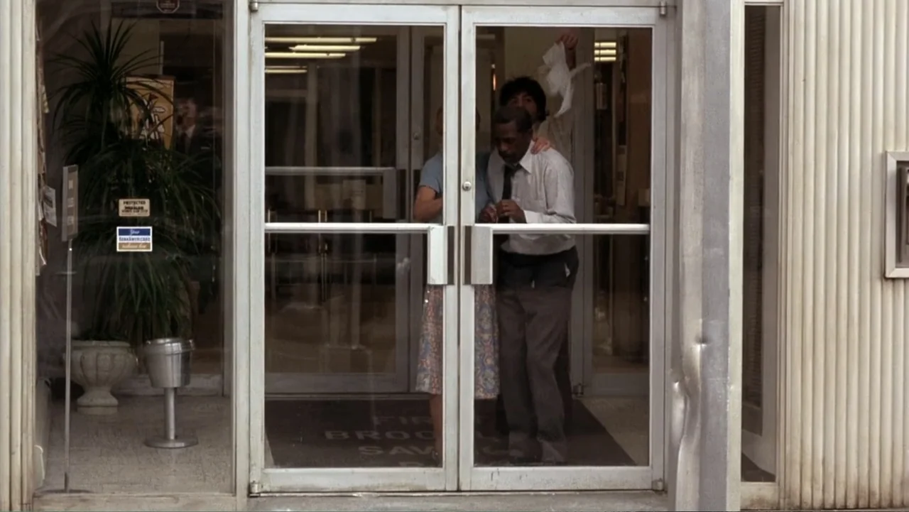 Dog Day Afternoon (1975) movie still: medium — A glass door bank entrance with a potted plant and a metal trash can. A man and a woman in 1970s…