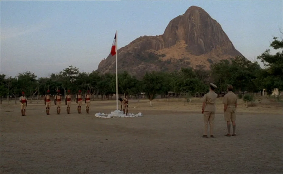 Chocolat (1988) movie still: wide — A dry, sandy parade ground sits at the base of a large rocky mountain. A row of soldiers stands at attention…