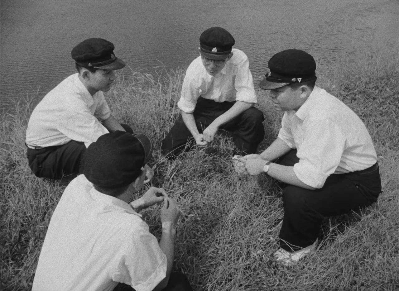 Hiroshima Mon Amour (1959) movie still: fullbody — Four school-aged boys crouch in a circle on a grassy bank near water. They are wearing…