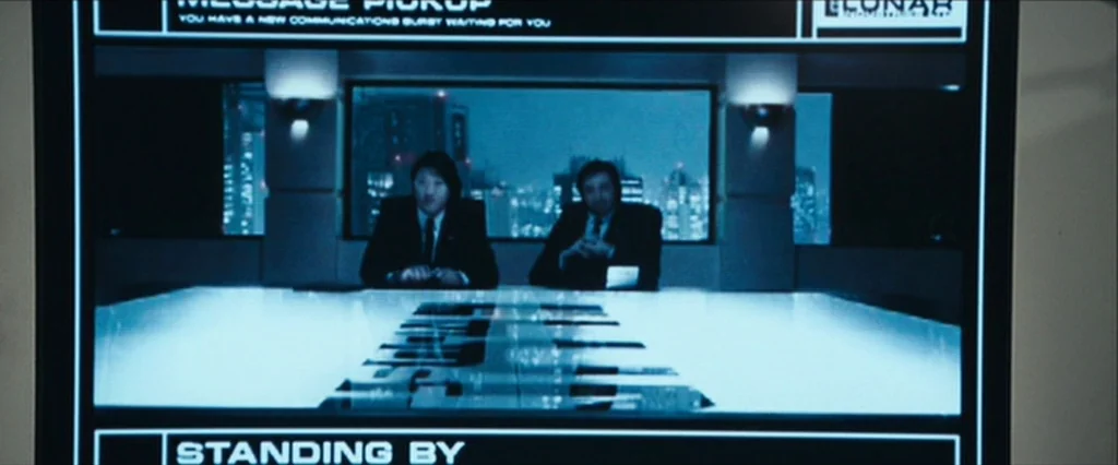 Moon (2009) movie still: medium — A digital computer screen displays a video transmission of two men in suits. The men sit at a glowing white…