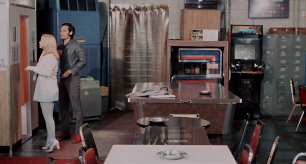 Buffalo ’66 (1998) movie still: In a dimly lit, wood-paneled corner of a 1970s arcade, a young couple pauses near a photo booth. The scene is framed…