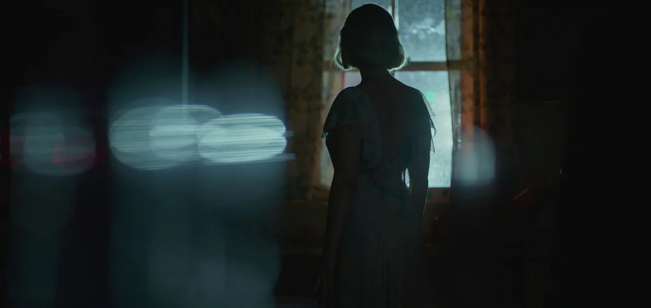 Foe (2023) movie still: medium — A dimly lit interior room at night with a single window providing light. A person with short hair stands facing the…