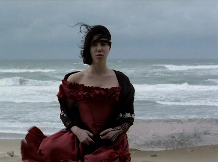 For Ever Mozart (1996) movie still: medium — A woman stands on a sandy beach under a grey, overcast sky. Waves crash in the background while wind…