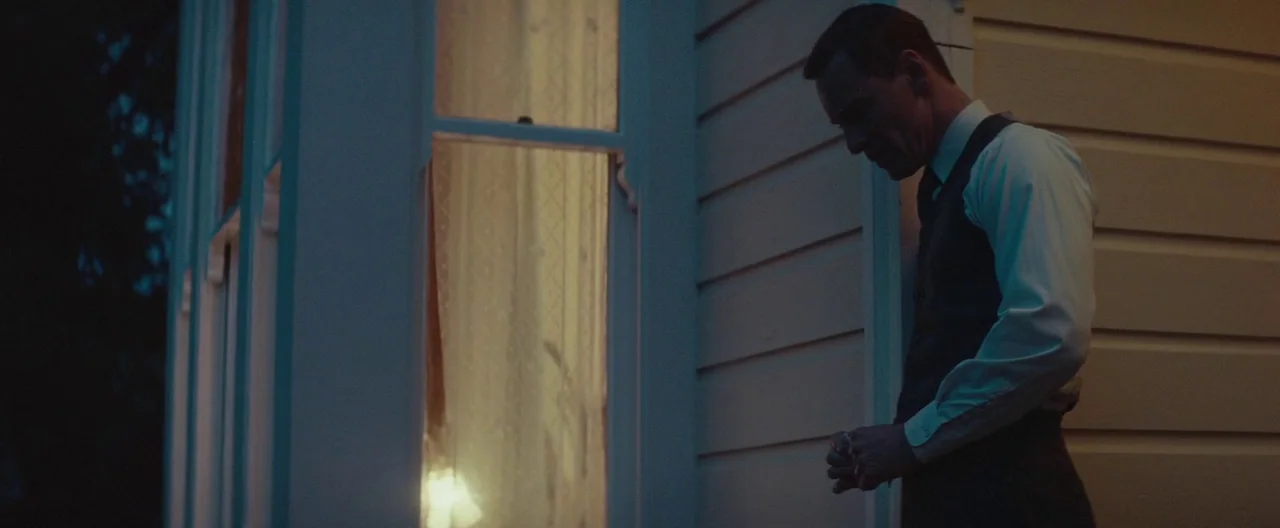 The Light Between Oceans (2016) movie still: medium — A man stands on a porch next to the illuminated window of a wooden house. He gazes downward…