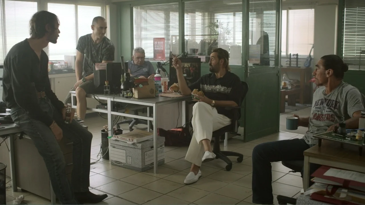 Unknown #1 movie still: fullbody — A utilitarian office space with glass partitions and tile flooring is filled with desks. Five men are gathered in…