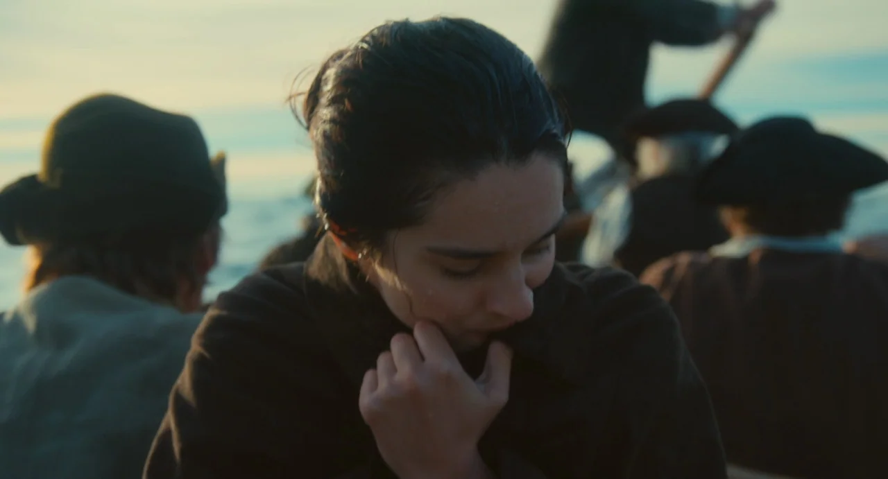 Portrait of a Lady on Fire movie still: closeup — Several people travel in a wooden boat upon an open body of water. A woman in a dark coat sits…