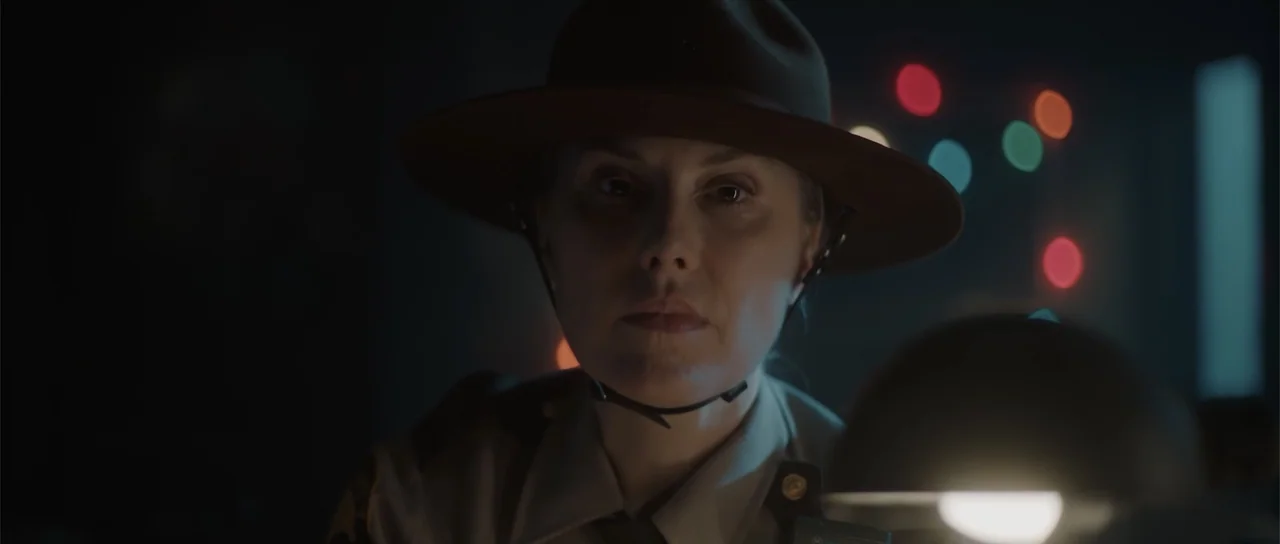 Night of the Missing (2023) movie still: closeup — A dimly lit room contains a police officer in uniform behind a desk. The officer sits facing the…