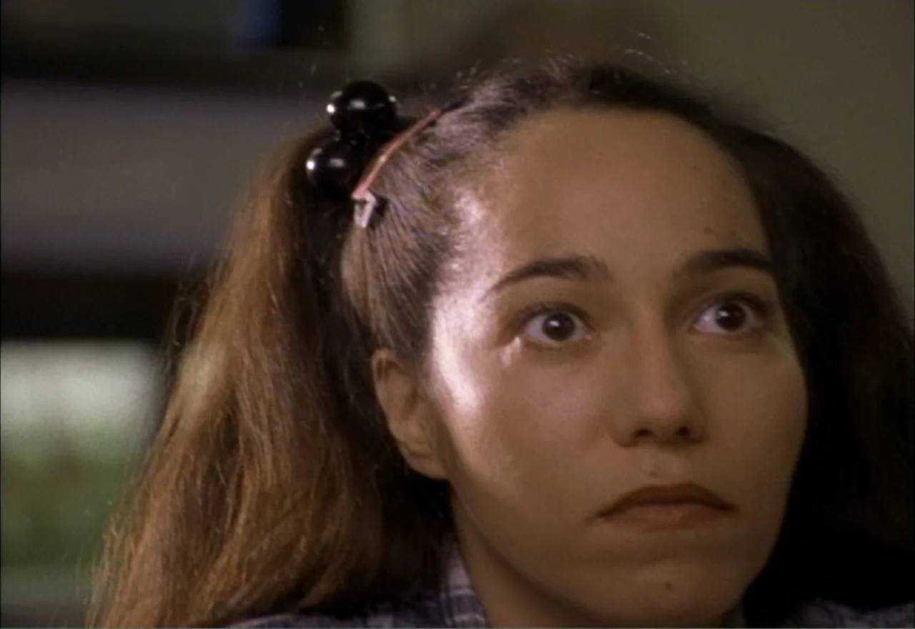 Ambushed (1998) movie still: closeup — An indoor space with indistinct neutral walls blurred in the background. A woman looks off-camera while her…
