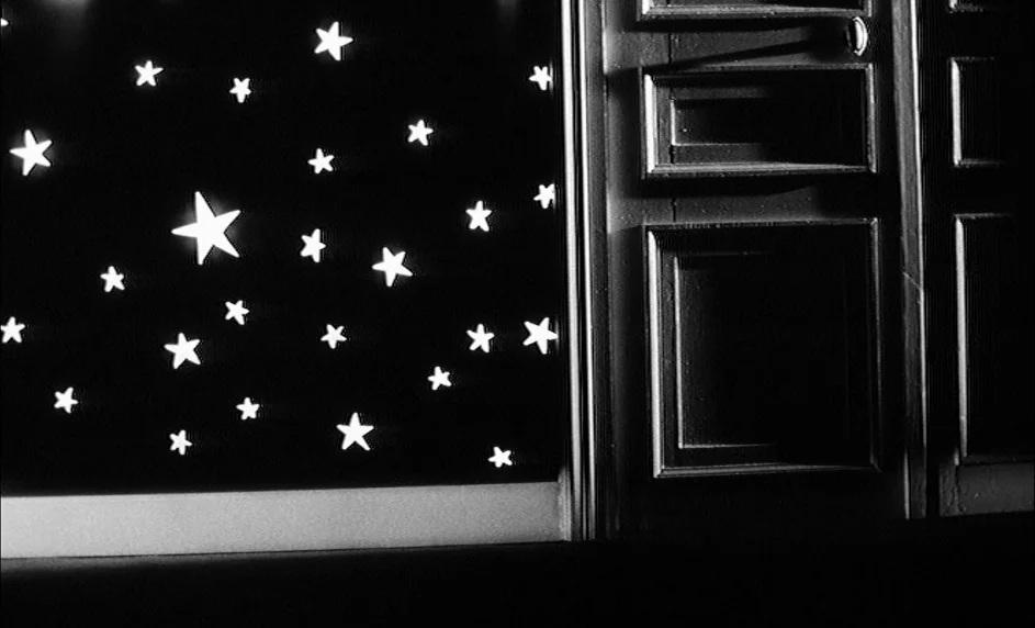 Boy Meets Girl (1984) movie still: wide — A dark wall is decorated with multiple cutout star shapes of varying sizes. Beside the wall, a tall, dark…
