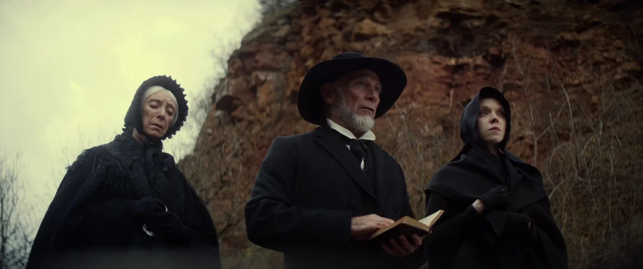 Never Grow Old (2019) movie still: medium — Three people in Victorian-era mourning attire stand before a rugged, natural orange-brown rock wall. The…