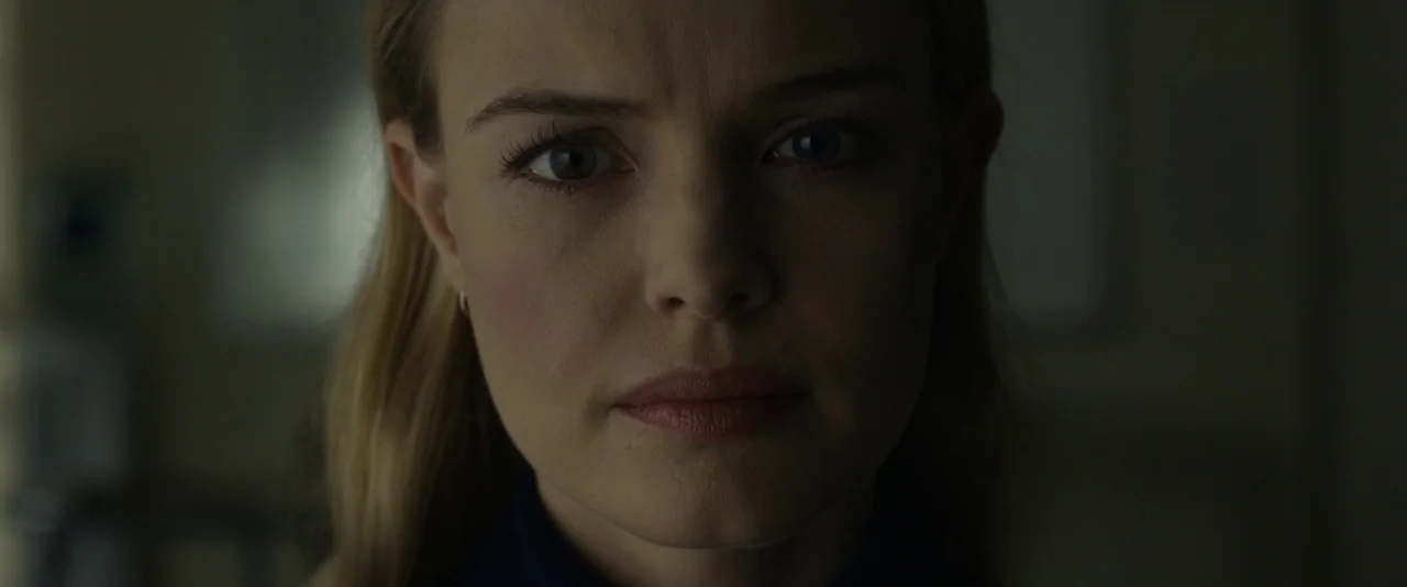Before I Wake (2016) movie still: closeup — A woman stands in a dimly lit, nondescript room with dark, shadowed surroundings. She faces the camera…