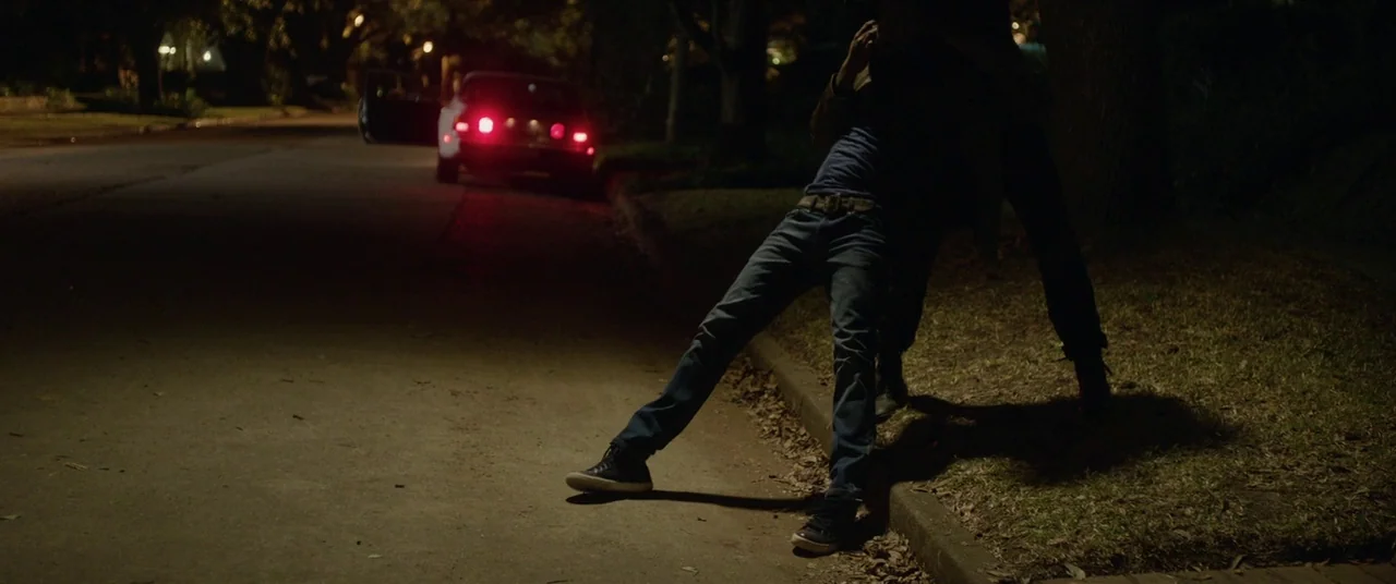 Get Out (2017) movie still: fullbody — A dark residential street at night features a concrete curb and overhanging dark tree branches. One person…