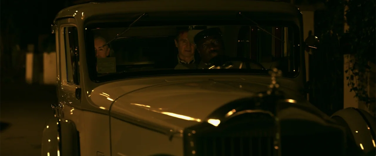 Marlowe (2023) movie still: medium — A vintage cream-colored sedan is parked on a dark street at night. Two men are seated in the front seats of the…