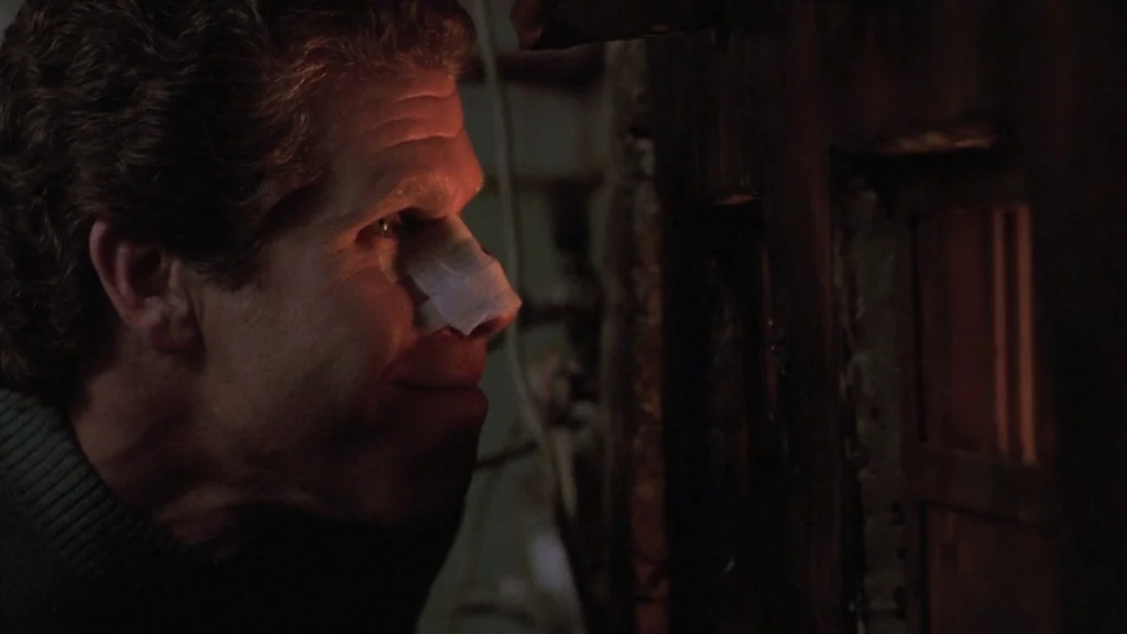 Cronos (1993) movie still: closeup — A man in a dark environment peers through a small rectangular cutout in a wall. The scene is illuminated by a…