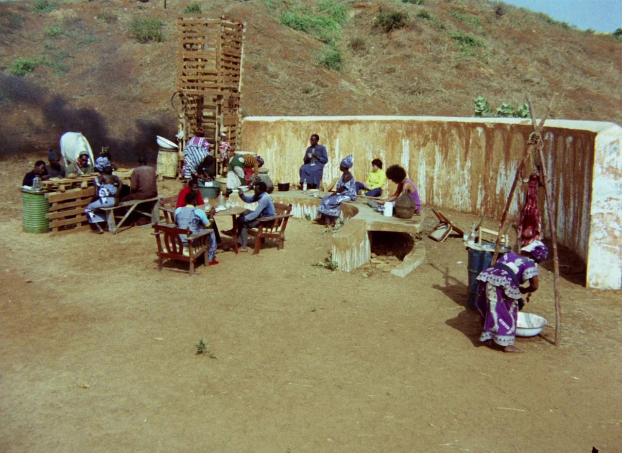 Touki Bouki (1973) movie still: wide — An open-air area with sandy ground, wooden structures, and a curved stone wall. People are seated at tables…