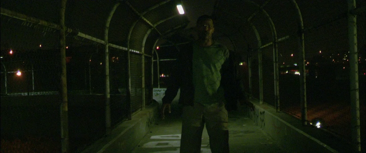 Collateral (2004) movie still: fullbody — A metal pedestrian overpass at night with chain-link fencing and concrete floors. A man stands in the…