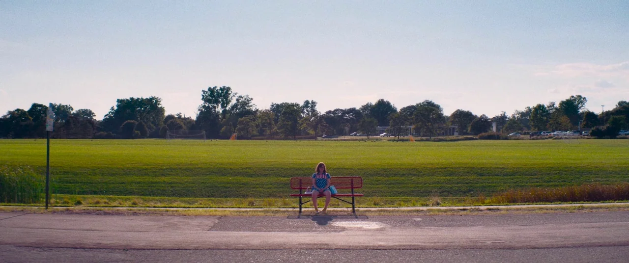Dinner in America (2020) movie still: wide — A wide expanse of mown green grass stretches toward a line of distant trees. A lone woman sits centered…