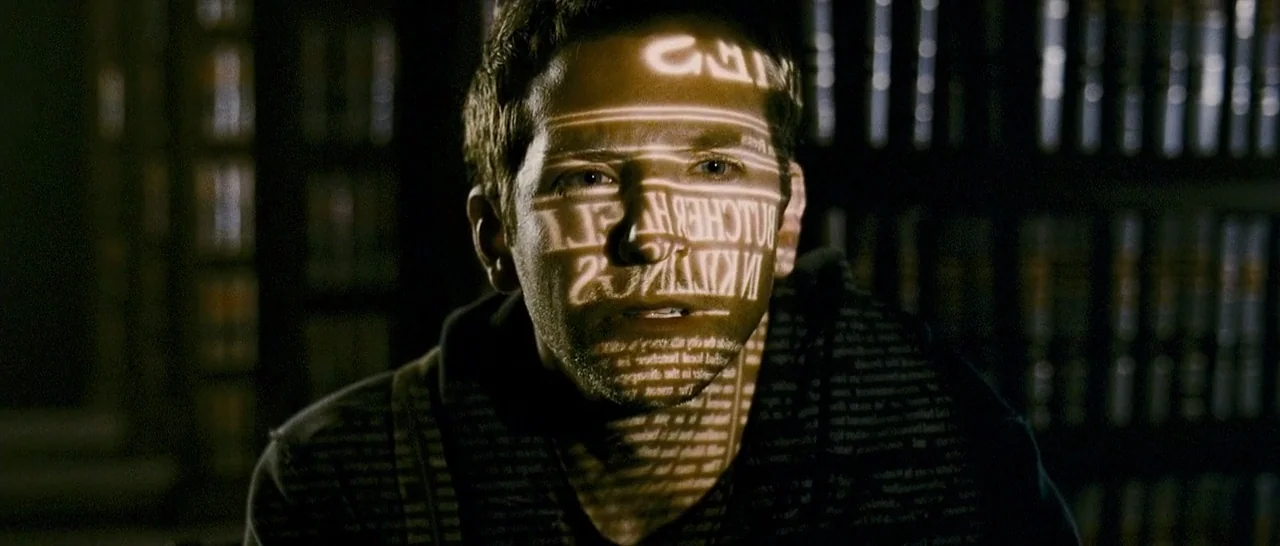 The Midnight Meat Train (2008) movie still: closeup — A dark home office with rows of books visible on shelves in the background. A man sits in…
