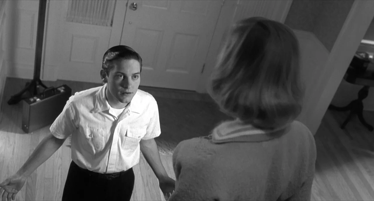 Pleasantville (1998) movie still: medium — A residential home entryway with a white door and wood flooring. A man stands facing a woman who has her…
