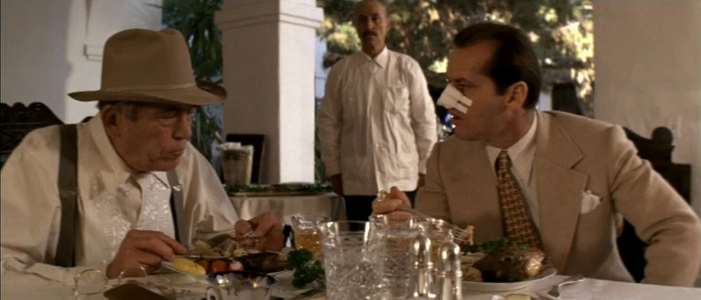 Chinatown (1974) movie still: medium — Two men sit at a white-clothed table eating a meal under a covered patio. A third man in a white shirt stands…