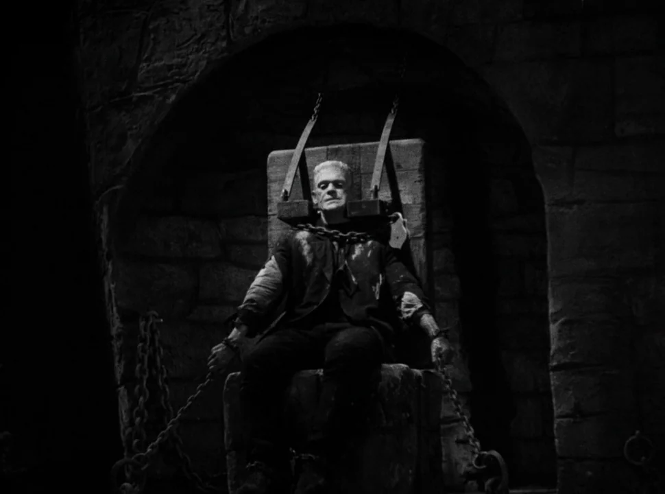 The Bride of Frankenstein (1935) movie still: fullbody — A stone alcove with textured blocks contains a man chained to a central block. He is bound…