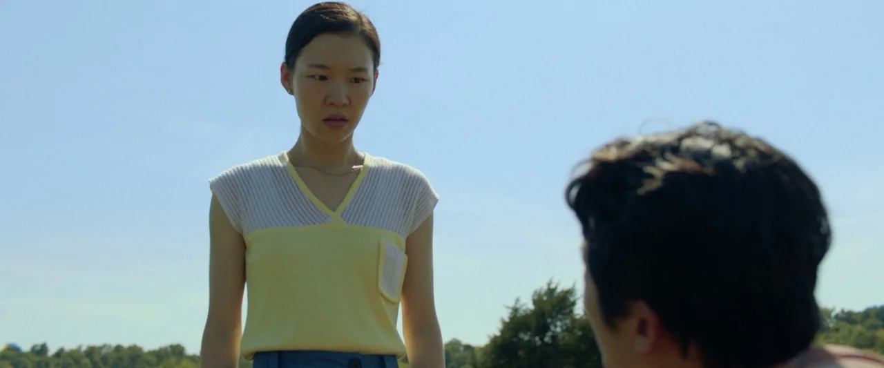 Minari (2020) movie still: medium — An open grassy field under a bright, cloudless blue sky. A woman stands looking downwards at an unseen person…