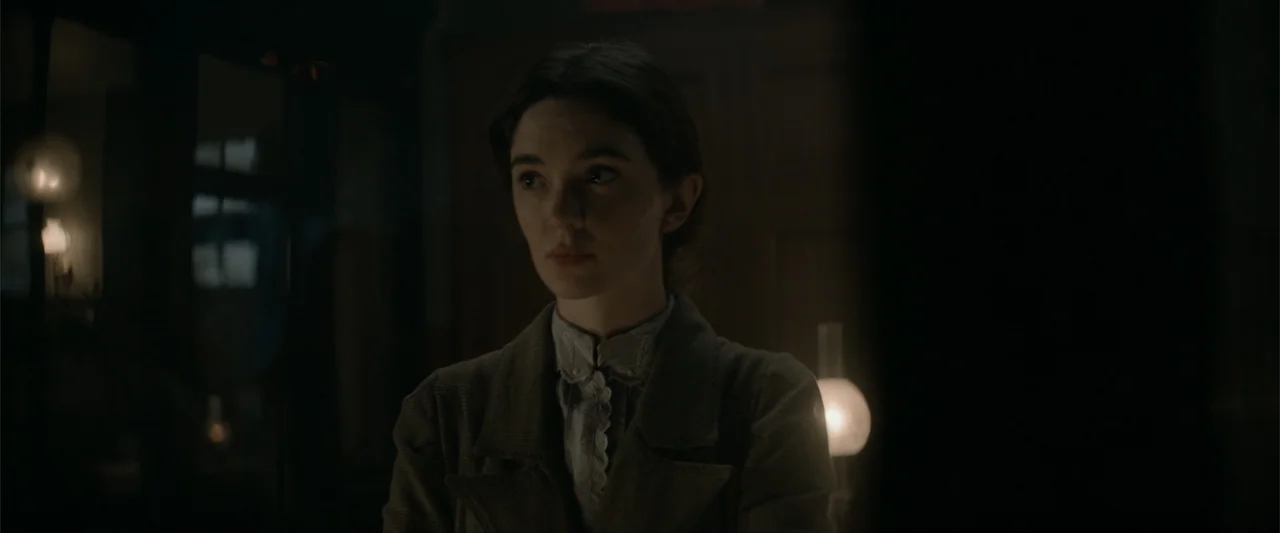 Eiffel (2021) movie still: medium — A dark, dimly lit room contains a young woman standing in the foreground. The subject looks slightly off-camera…