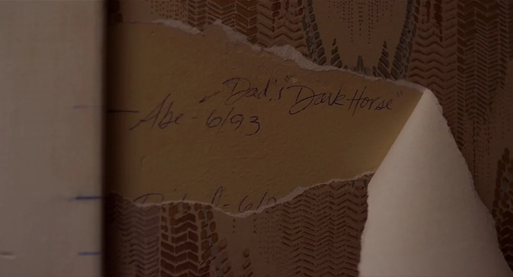 Dark Horse (2011) movie still: closeup — A section of wall is revealed where old wallpaper has been peeled away. Handwritten notes are visible on…