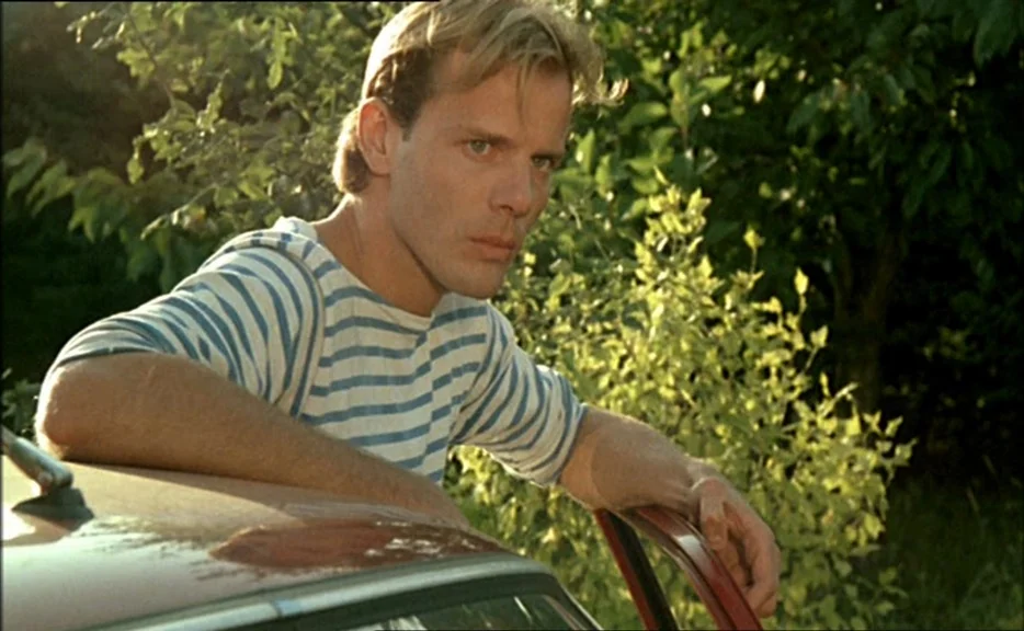 Pauline At the Beach (Pauline à la plage) (1983) movie still: medium — A man stands outdoors near trees while leaning against the roof of a car. He…