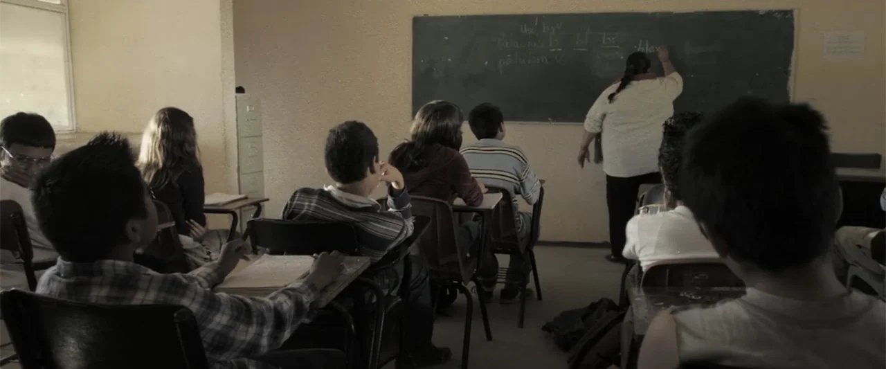 At Dawn (2013) movie still: medium — A sparsely furnished classroom features plain beige walls, wooden desks, and a dark blackboard. An instructor…