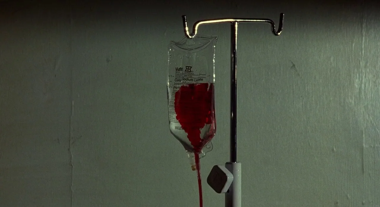 Hellraiser (1987) movie still: closeup — A medical IV fluid bag filled with red liquid hangs on a metal pole. The stand is positioned against a…