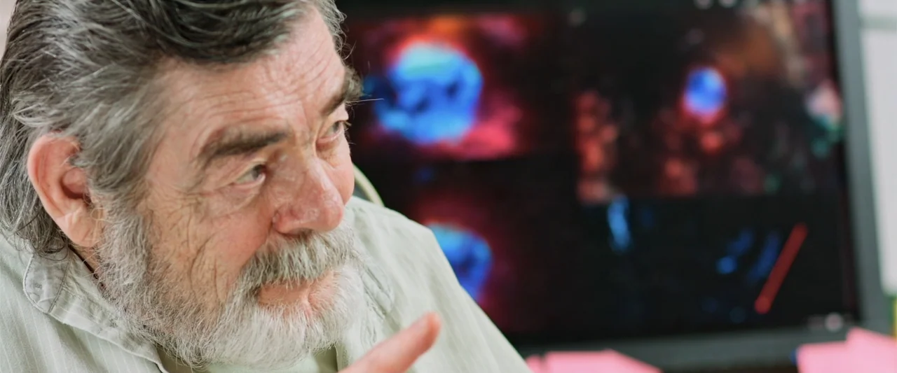 Beyond the Universe (2022) movie still: closeup — An office interior features an elderly man sitting in front of a computer monitor. The man points…