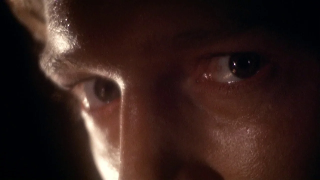 The Day of the Locust (1975) movie still: closeup — A man's eyes stare intently from the darkness. The face is partially lit by a harsh light source…