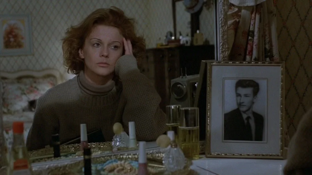 Magic (1978) movie still: medium — A woman sits at a vanity with patterned wallpaper and an ornate mirror. She gazes toward a framed black-and-white…