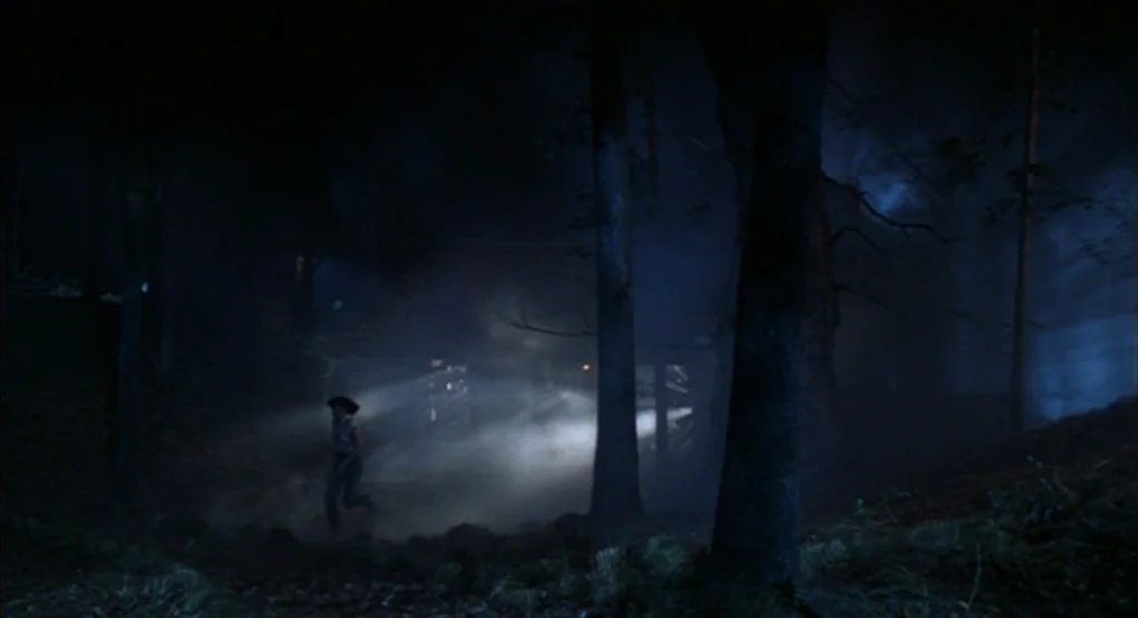 Evil Dead II (1987) movie still: wide — A dark, dense woodland area is filled with thick ground fog at night. A woman runs through the haze while…