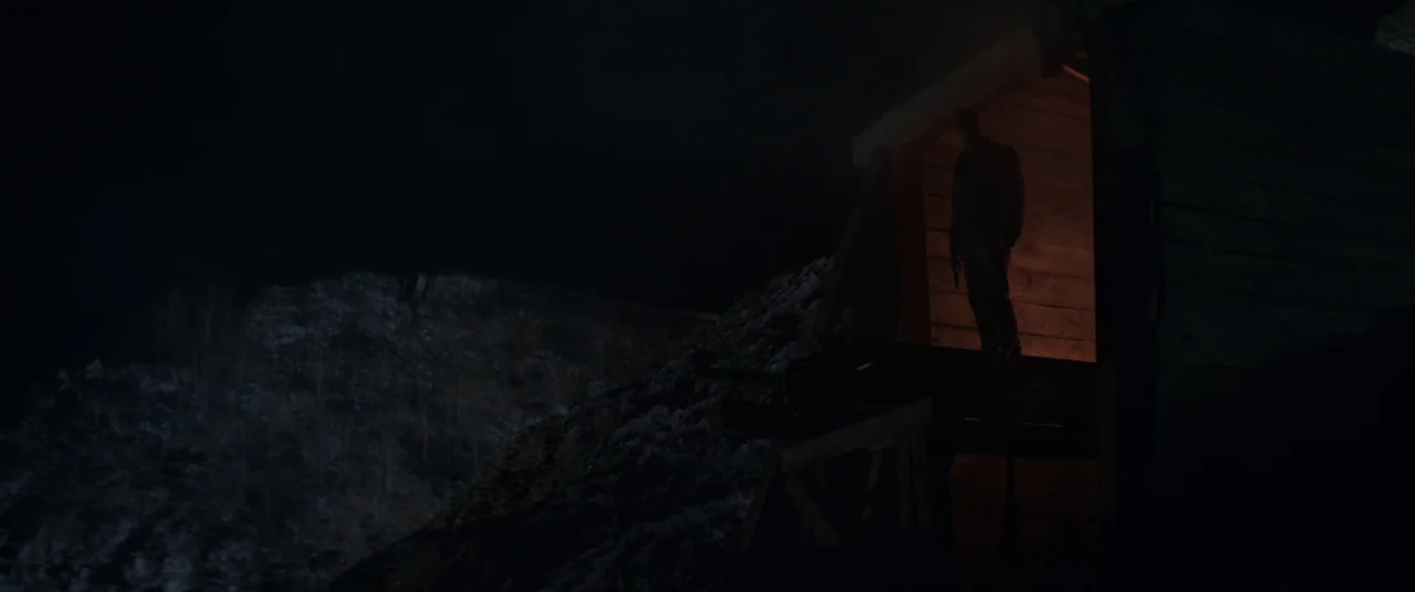 Hold The Dark (2018) movie still: wide — A dark, snow-dusted hillside at night features a small illuminated wooden entrance structure. A man stands…