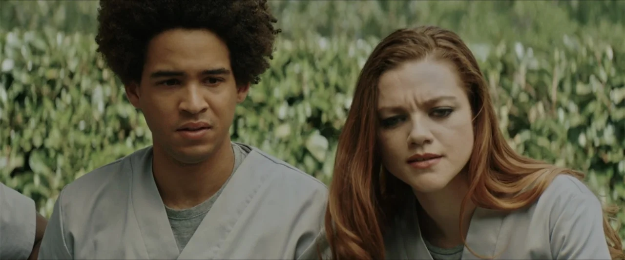 American Carnage (2022) movie still: medium — Two young adults stand outdoors against a blurred backdrop of dense green hedges. They are wearing…