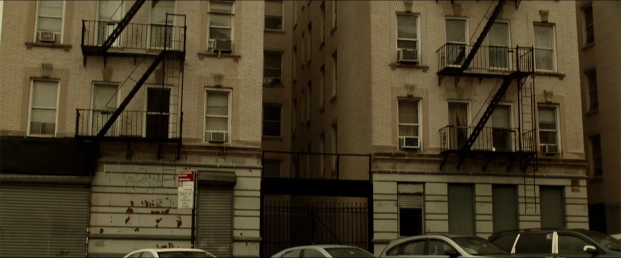 Exposed (2016) movie still: wide — A dense urban alleyway is flanked by two multi-story brick apartment buildings. Iron fire escapes are attached to…