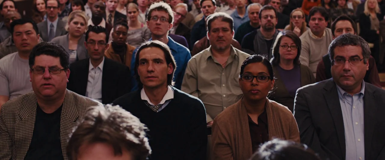 The Wolf of Wall Street (2013) movie still: medium — A large crowd sits in rows inside a crowded conference room. They face forward in quiet…